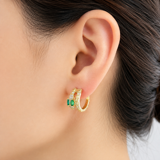 A close-up of a person's ear wearing a small gold huggie hoop earring with green rectangular gemstones and small clear stones.
