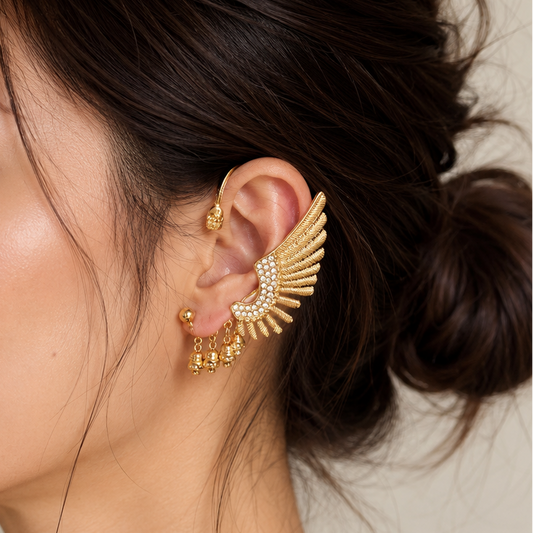 A woman in profile wearing a gold wing-shaped ear cuff earring with small clear stones and dangling gold charms, against a soft beige background.