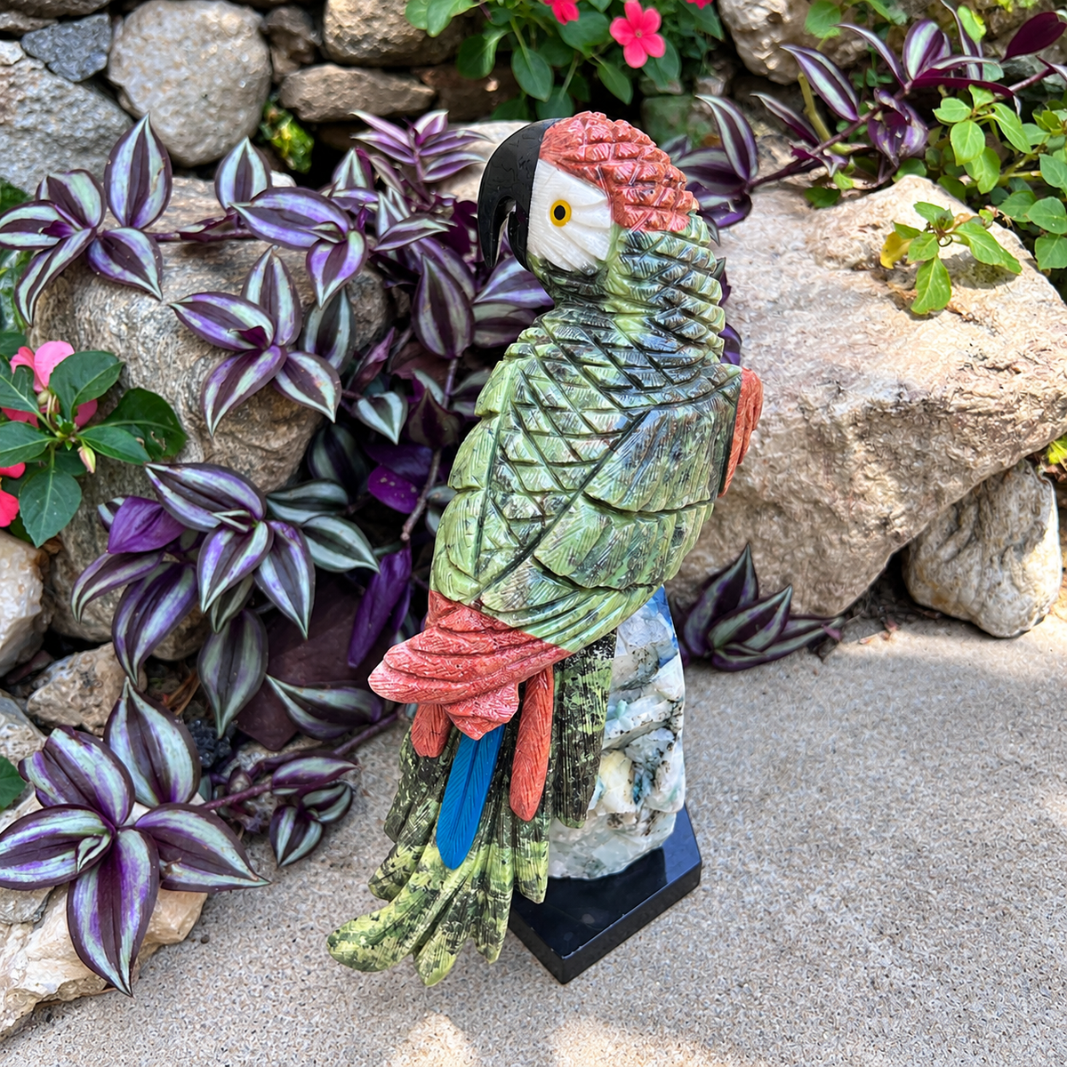 A decorative parrot garden statue perched on a rock, styled outdoors among purple and green trailing plants and natural stones.