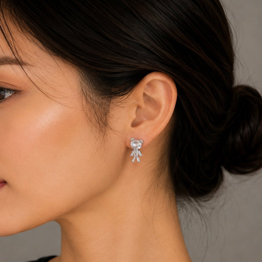 A close-up of a woman's ear wearing a small silver teddy bear stud earring with clear stones, set against a neutral gray background.