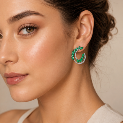 A close-up of a woman wearing a crescent-shaped dangle earring with green stones and a silver outer ring set with small clear stones, against a soft beige background.