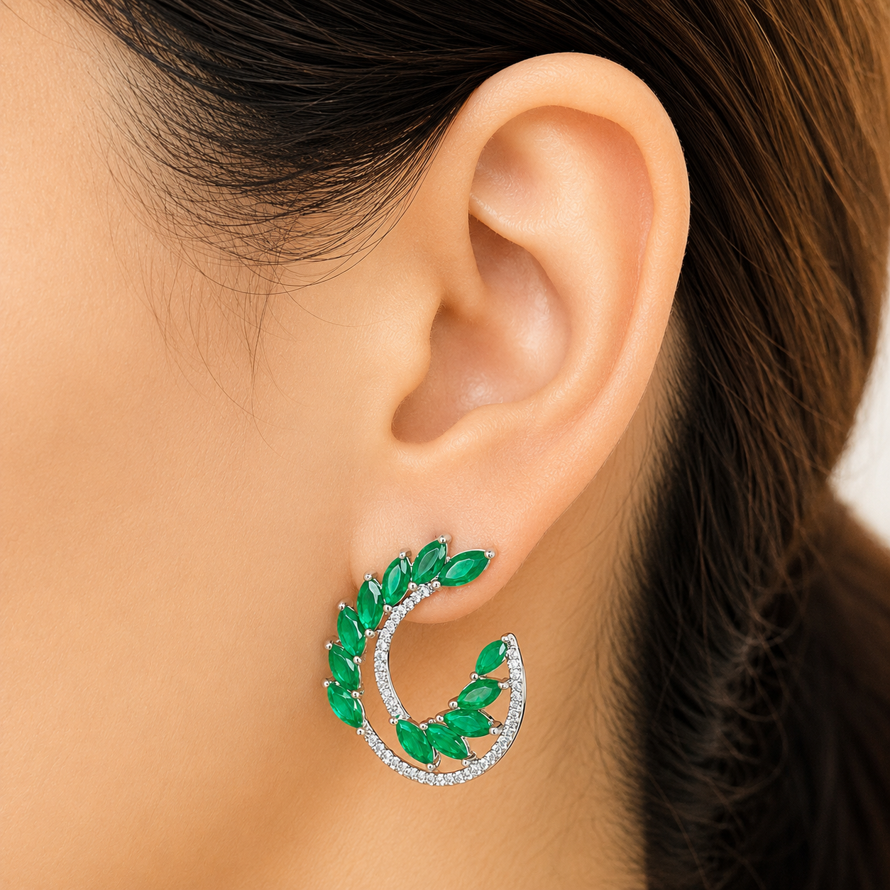 A close-up of a model's ear wearing a crescent-shaped hoop earring with green marquise-cut stones and a silver inner hoop set with small clear stones.