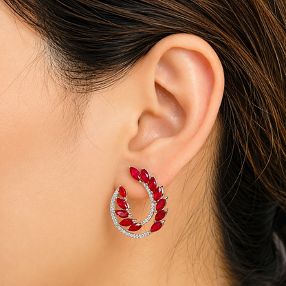 A close-up of a model's ear wearing a silver crescent-shaped hoop earring adorned with red teardrop crystals and a border of small clear stones.