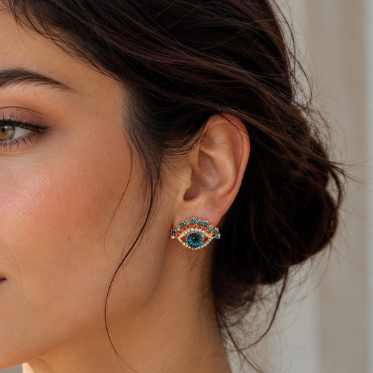 Close-up of a woman wearing a small eye-shaped stud earring with turquoise blue, red, and gold details, shown in warm natural light.