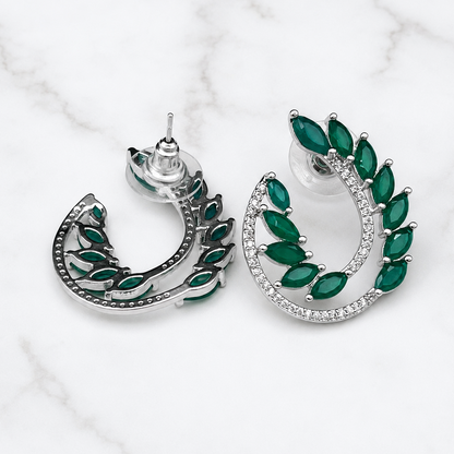 A pair of silver crescent-shaped earrings with marquise-cut green gemstones and a row of clear stones, displayed on a white marble background.