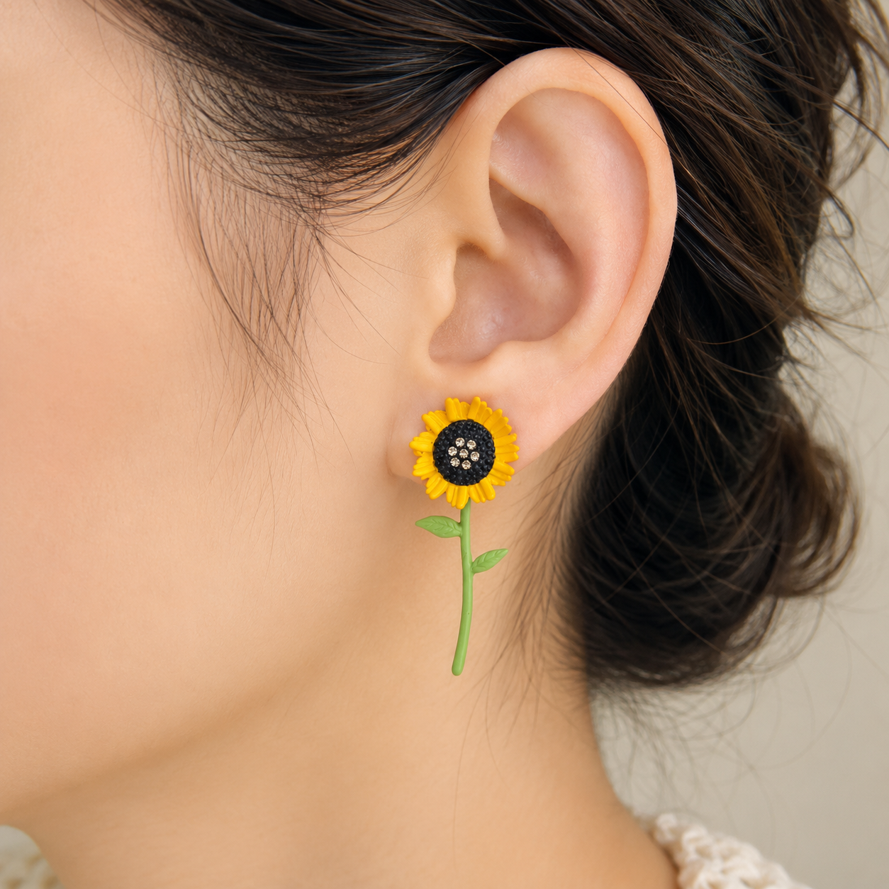 A close-up of a person's ear wearing a small sunflower stud earring with yellow petals, a dark center, and a green stem against a soft light beige background.