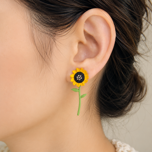 A close-up of a person's ear wearing a small sunflower stud earring with yellow petals, a dark center, and a green stem against a soft light beige background.