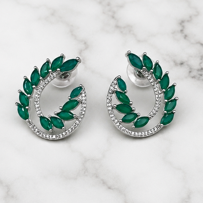 A pair of silver crescent-shaped hoop earrings with marquise-cut green gemstones and a border of small clear stones, displayed on a white marble background.