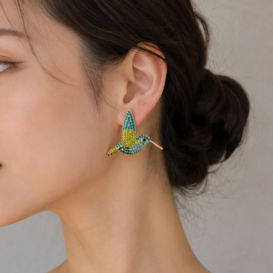 A woman in a black spaghetti-strap top wearing a gold and turquoise hummingbird earring, shown in a close-up profile against a neutral gray background.
