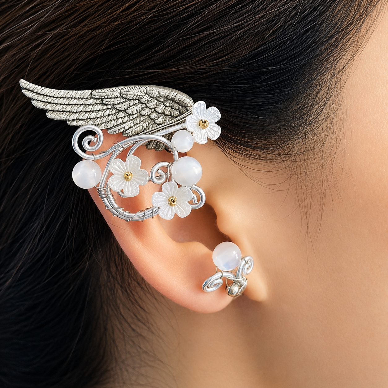 A close-up of a person's ear wearing an ornate silver wing and floral ear cuff with white flower charms and pearl-like beads, plus a matching small hoop earring.