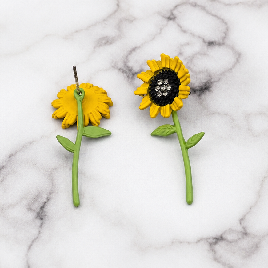 A pair of yellow sunflower stud earrings with green stems and leaves, displayed on a white and gray marble surface.