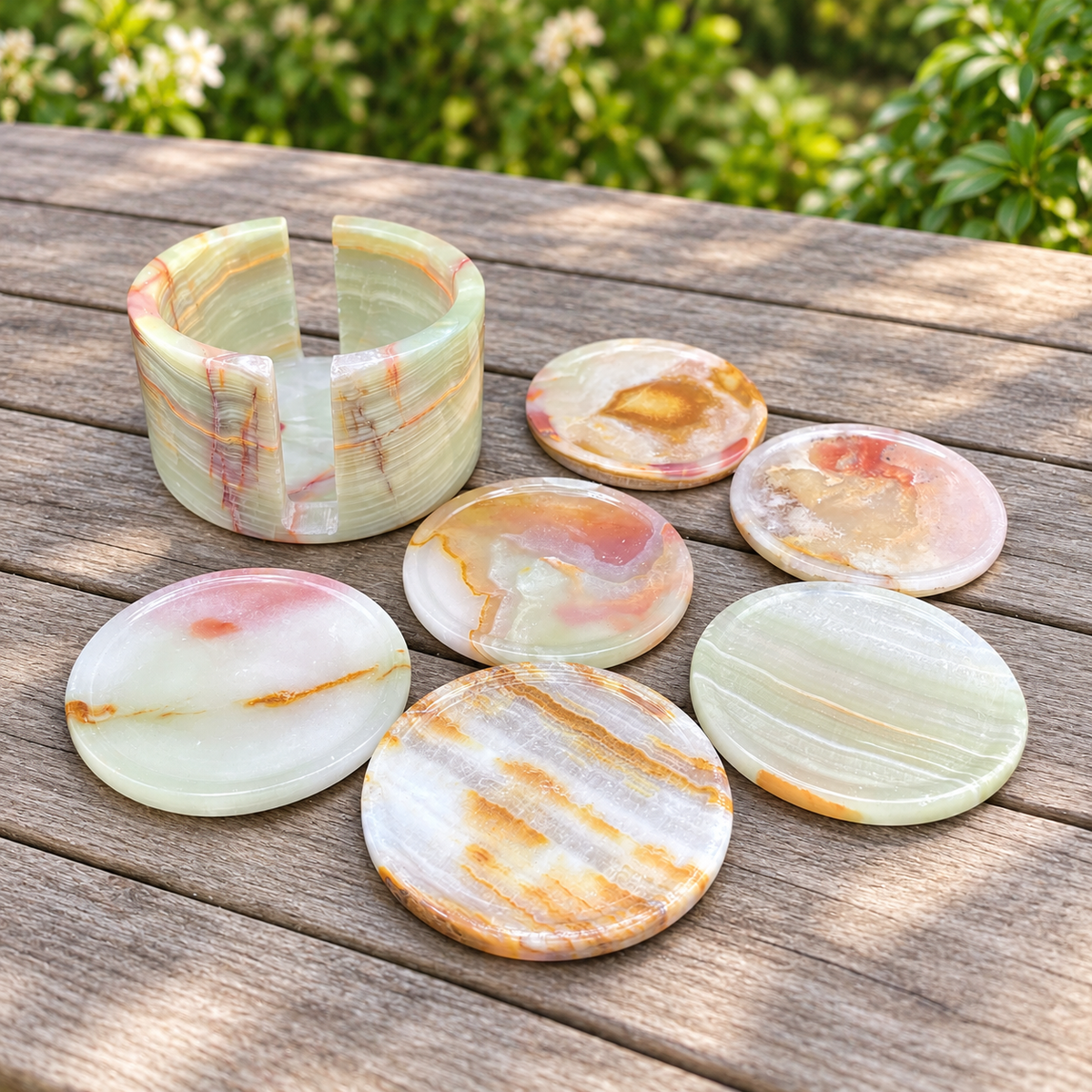 A set of carved stone coasters and a matching cylindrical tray with a wide band, displayed on a weathered wooden table outdoors with green foliage in the background.
