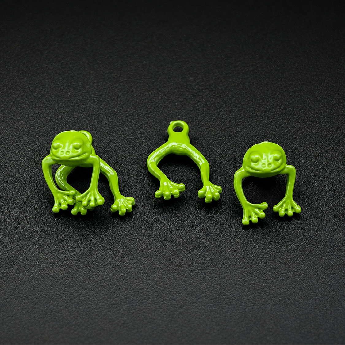 Three small bright green frog-shaped charms or pendants arranged on a dark textured background. Each frog has a loop at the top, suggesting they are jewelry components or accessories.