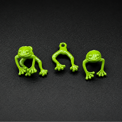 Three small bright green frog-shaped charms or pendants arranged on a dark textured background. Each frog has a loop at the top, suggesting they are jewelry components or accessories.