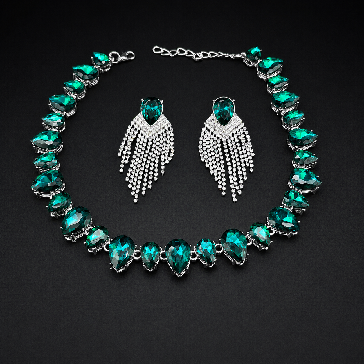 A jewelry set featuring a silver-tone necklace and matching drop earrings with emerald-cut green crystals and silver pavé fringe details, displayed on a dark background.