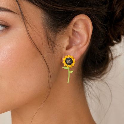 A close-up of a woman's ear wearing a small sunflower stud earring with a yellow petal, black center, and green stem against a soft beige background.