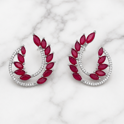 A pair of silver crescent-shaped earrings with marquise-cut red gemstones and rows of small clear stones, displayed on a white marble surface.