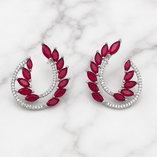 A pair of silver crescent-shaped earrings with marquise-cut red gemstones and rows of small clear stones, displayed on a white marble surface.