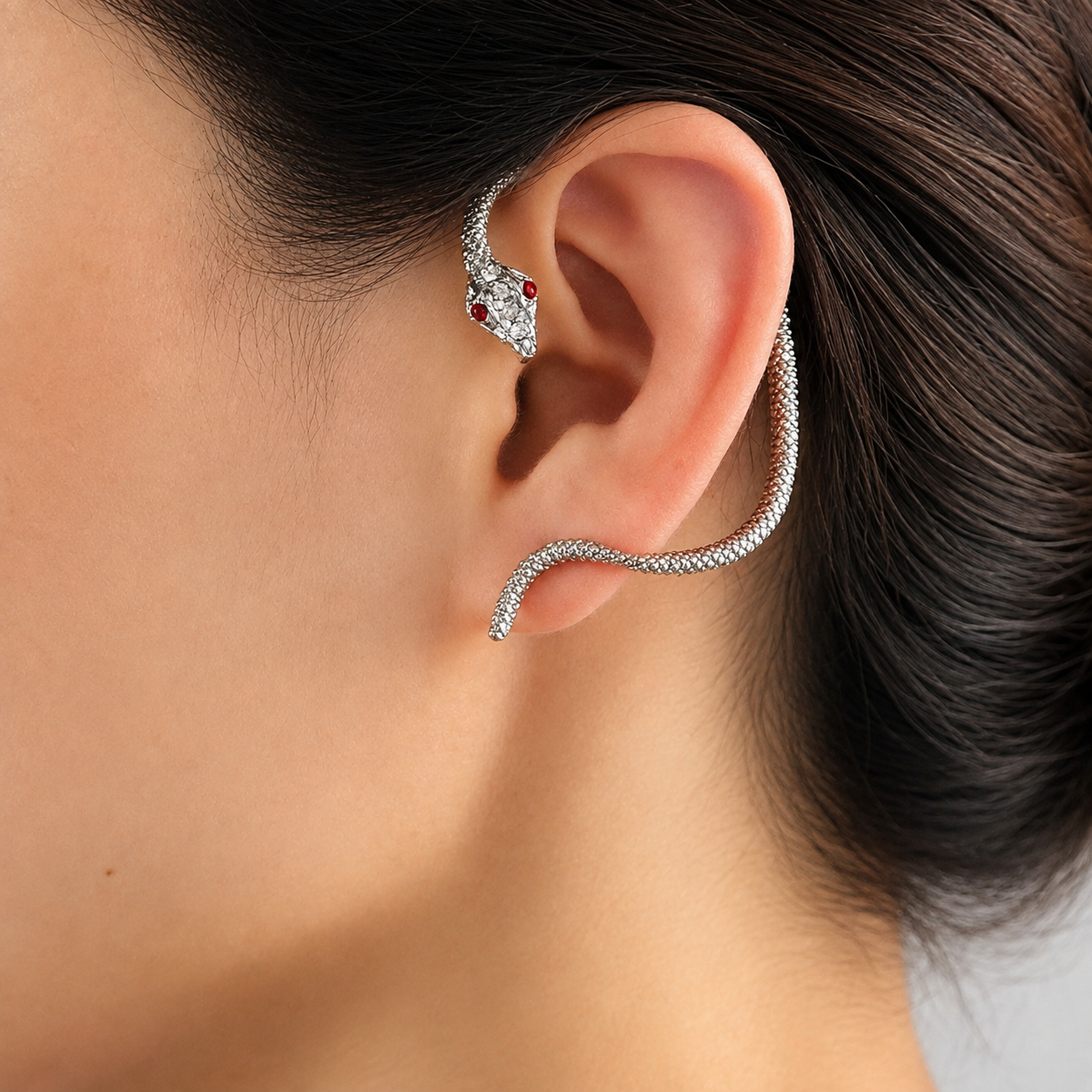 A close-up of a person's ear wearing a silver snake-shaped ear cuff earring with small red gemstone accents on the snake's head.