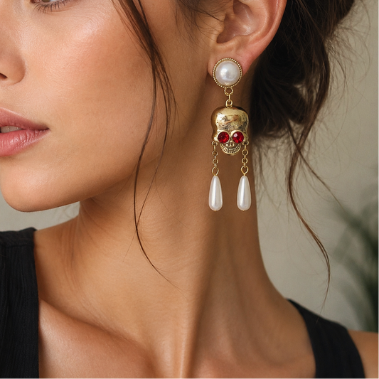 A close-up of a woman wearing a dangling gold skull earring with red gemstone eyes and white pearl drops, shown against a soft neutral background.