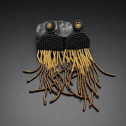 A pair of long beaded fringe earrings displayed on a dark textured stand against a dark gray background. The earrings feature black and gold seed beads in a tiered arch design with long dangling fringe.