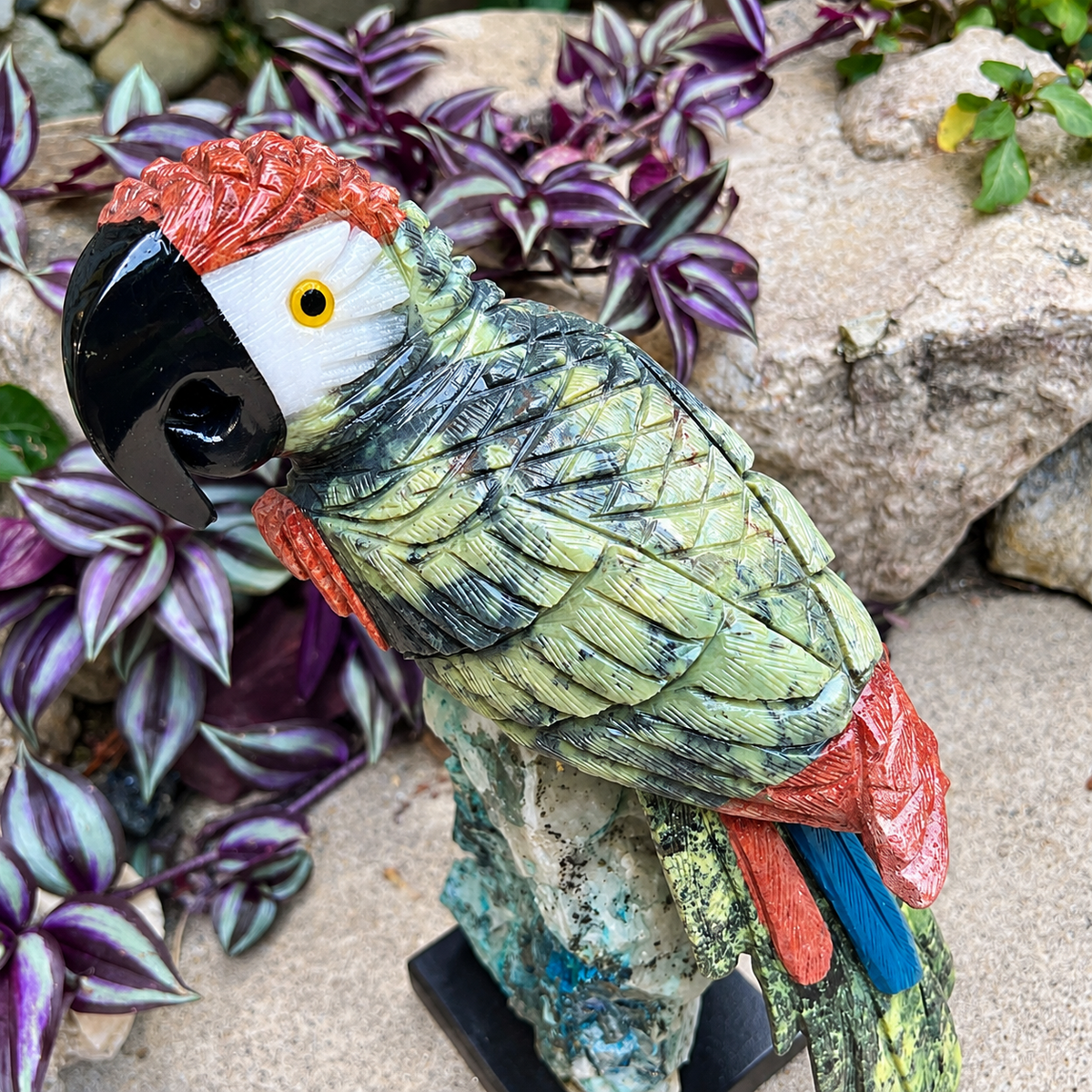 A decorative parrot garden statue with textured green, black, red, and blue feathers, perched on a small stone-like base in a garden setting with purple foliage and rocks.