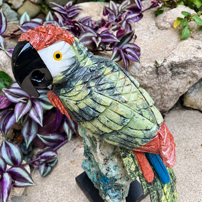 A decorative parrot garden statue with textured green, black, red, and blue feathers, perched on a small stone-like base in a garden setting with purple foliage and rocks.