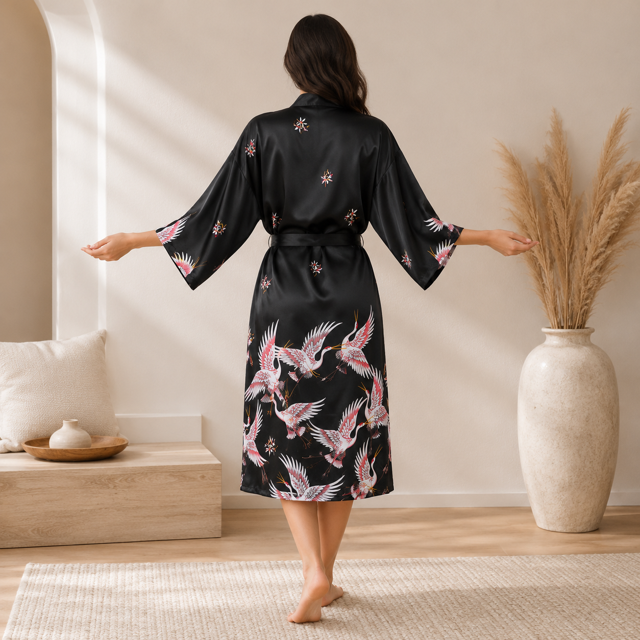 A woman stands barefoot indoors wearing a black satin-like robe with pink and white crane prints and small floral motifs. The robe has wide sleeves and a self-tie waist, shown from the back in a softly lit room with a bench, pillows, and a large vase of dried grass.