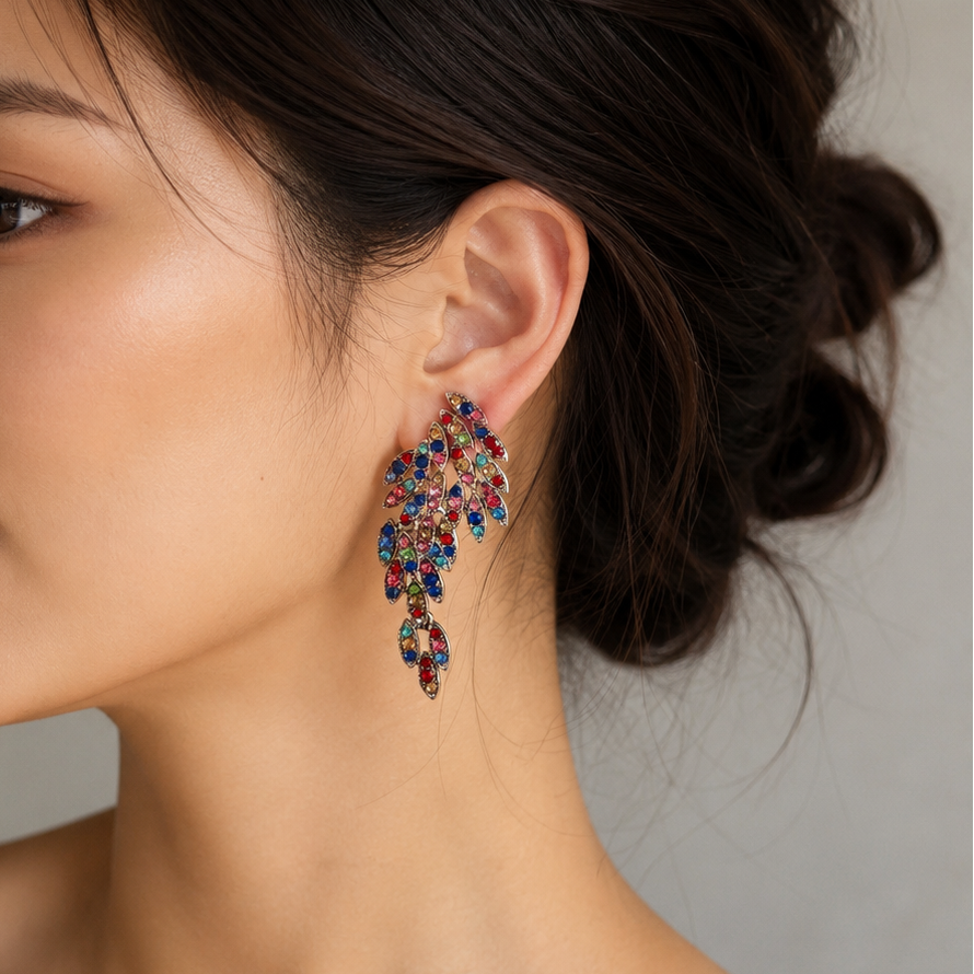 A close-up side profile of a woman wearing a large, colorful dangling earring with multiple faceted gemstones in blue, red, pink, and gold-toned metal, against a soft gray background.