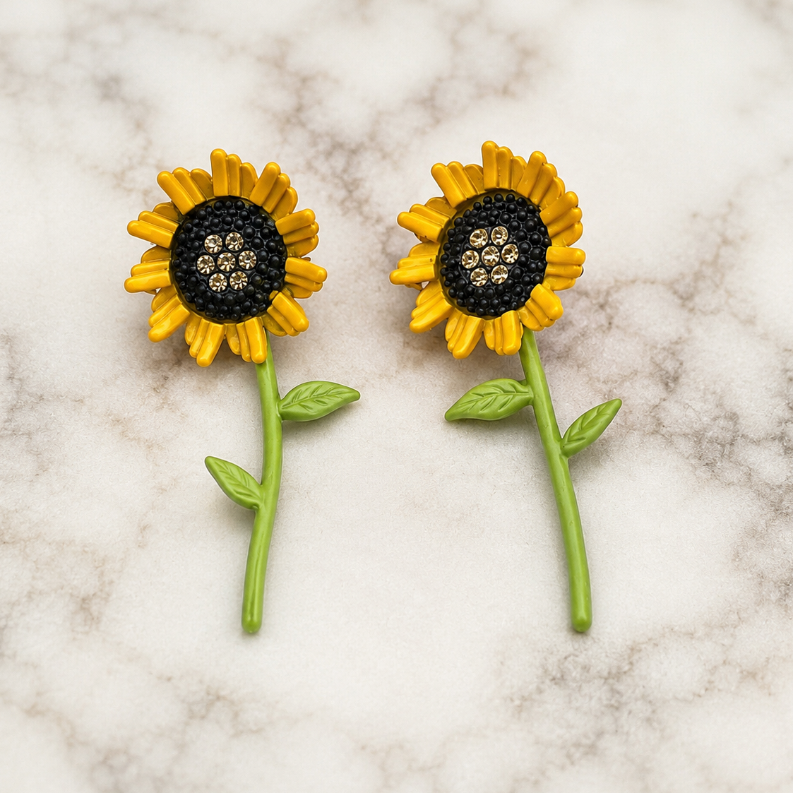 A pair of sunflower-shaped earrings with yellow petals, black flower centers, and green stems, displayed on a white and gray marble surface.