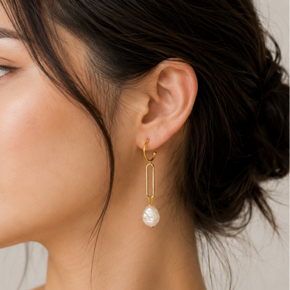 A close-up side profile of a woman wearing a delicate gold huggie hoop earring with a dangling bar and a single white freshwater pearl drop, set against a soft neutral background.