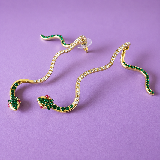 Gold snake-shaped dangle earrings embellished with green rhinestones and small red rhinestones, displayed on a solid purple background.