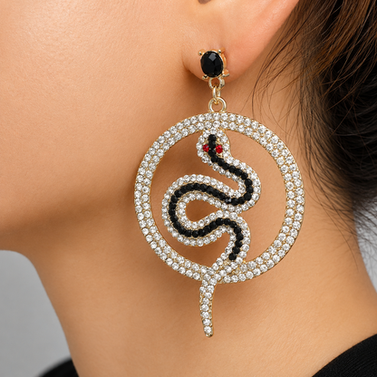 A close-up of a person's ear wearing a large snake-shaped earring. The earring features a gold-tone setting, a circular border paved with clear rhinestones, a black snake body with a small red eye, and a rhinestone-encrusted tail.