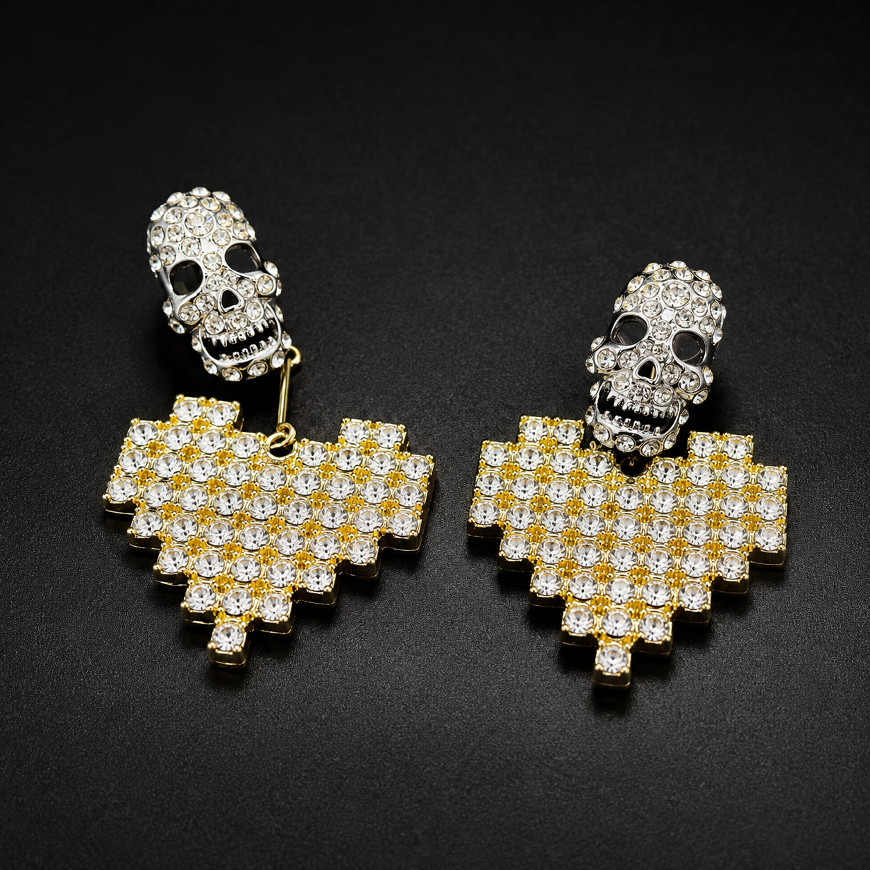 A pair of gold and silver skull drop earrings adorned with clear rhinestones, displayed on a dark textured background.
