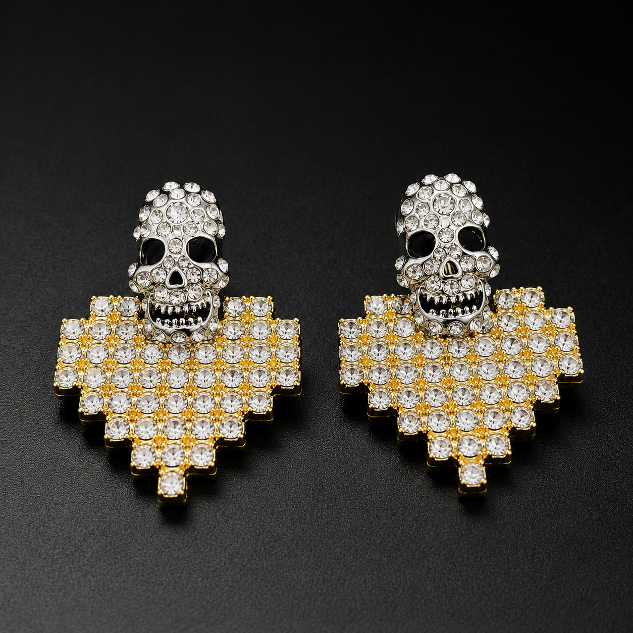 A pair of gothic skull stud earrings with silver-toned skulls and gold-toned pixelated heart-shaped bases paved with clear rhinestones, set against a dark textured background.