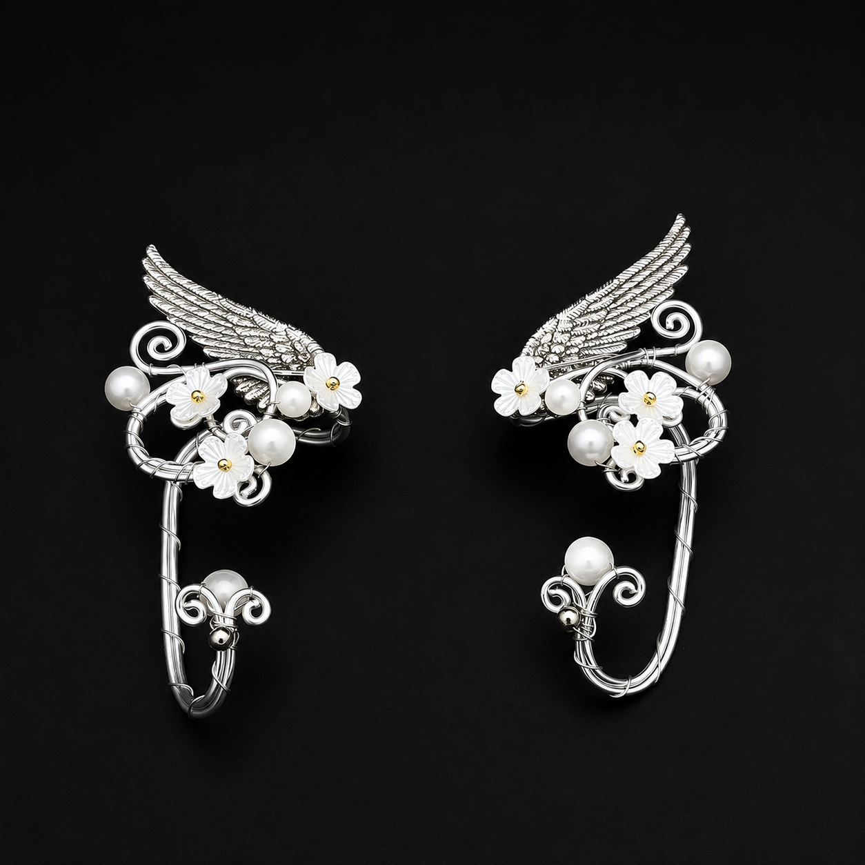 A pair of silver-toned winged hoop earrings decorated with white cherry blossom flowers and pearl-like beads, displayed on a solid black background.