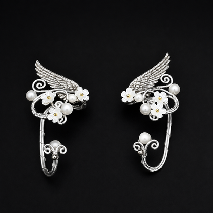 A pair of silver-toned winged hoop earrings decorated with white cherry blossom flowers and pearl-like beads, displayed on a solid black background.