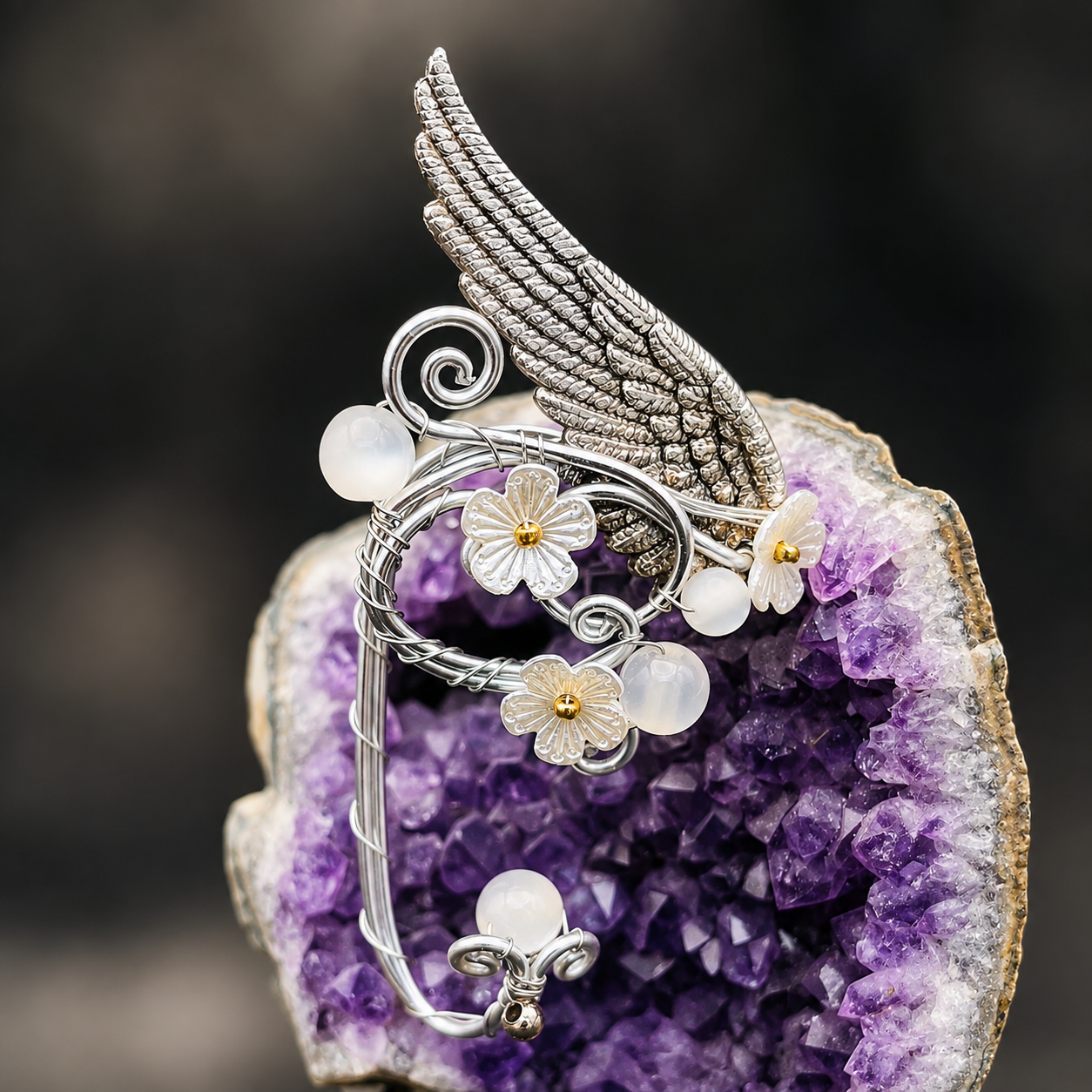 A silver-toned winged wire art piece with white flower charms and iridescent pearl-like beads, displayed on a purple crystal geode against a dark blurred background.