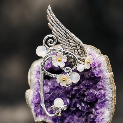 A silver-toned winged wire art piece with white flower charms and iridescent pearl-like beads, displayed on a purple crystal geode against a dark blurred background.