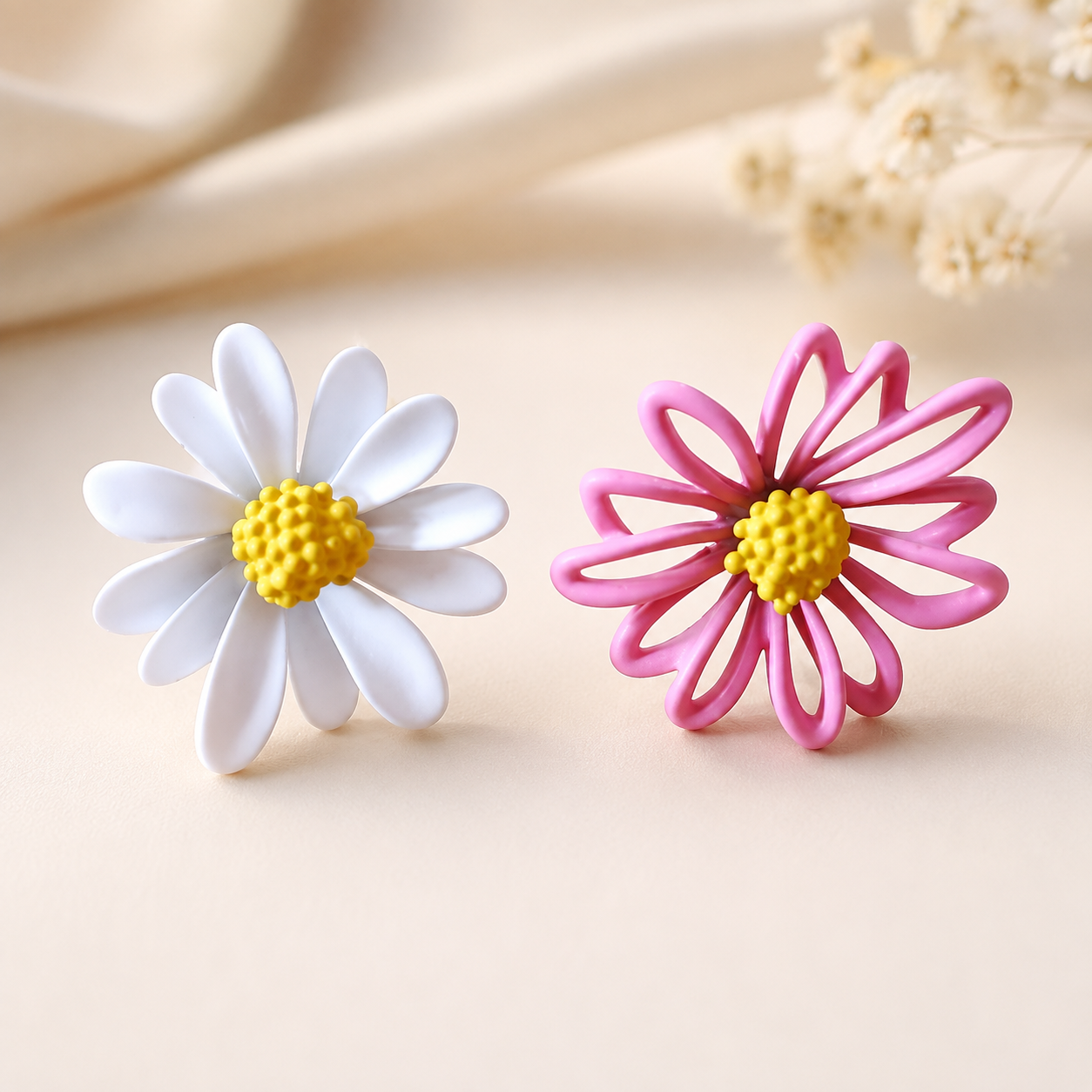 Two daisy-shaped earrings on a warm beige surface, one with white petals and one with pink petals, both with yellow centers.