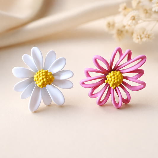 Two daisy-shaped earrings on a warm beige surface, one with white petals and one with pink petals, both with yellow centers.