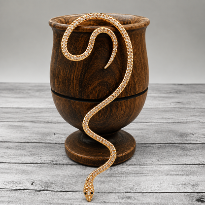 A gold snake figurine draped over a dark brown wooden cup, set on a gray wooden plank surface against a neutral gray background.