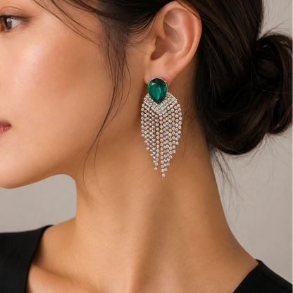 A close-up side profile of a woman wearing a dramatic statement dangle earring. The earring features a teardrop-shaped green emerald-cut stone at the top, set in silver-toned metal and adorned with cascading rows of clear pavé-set stones.
