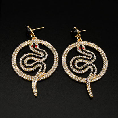 A pair of gold-tone snake drop earrings paved with clear rhinestones, featuring small red gemstone eyes and black posts, set against a solid black background.