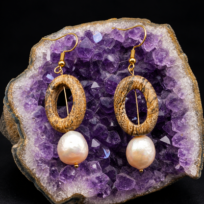 A pair of dangling earrings displayed on a purple crystal geode. Each earring has a textured natural wood oval frame, a gold-tone wire center, and a dangling freshwater pearl.