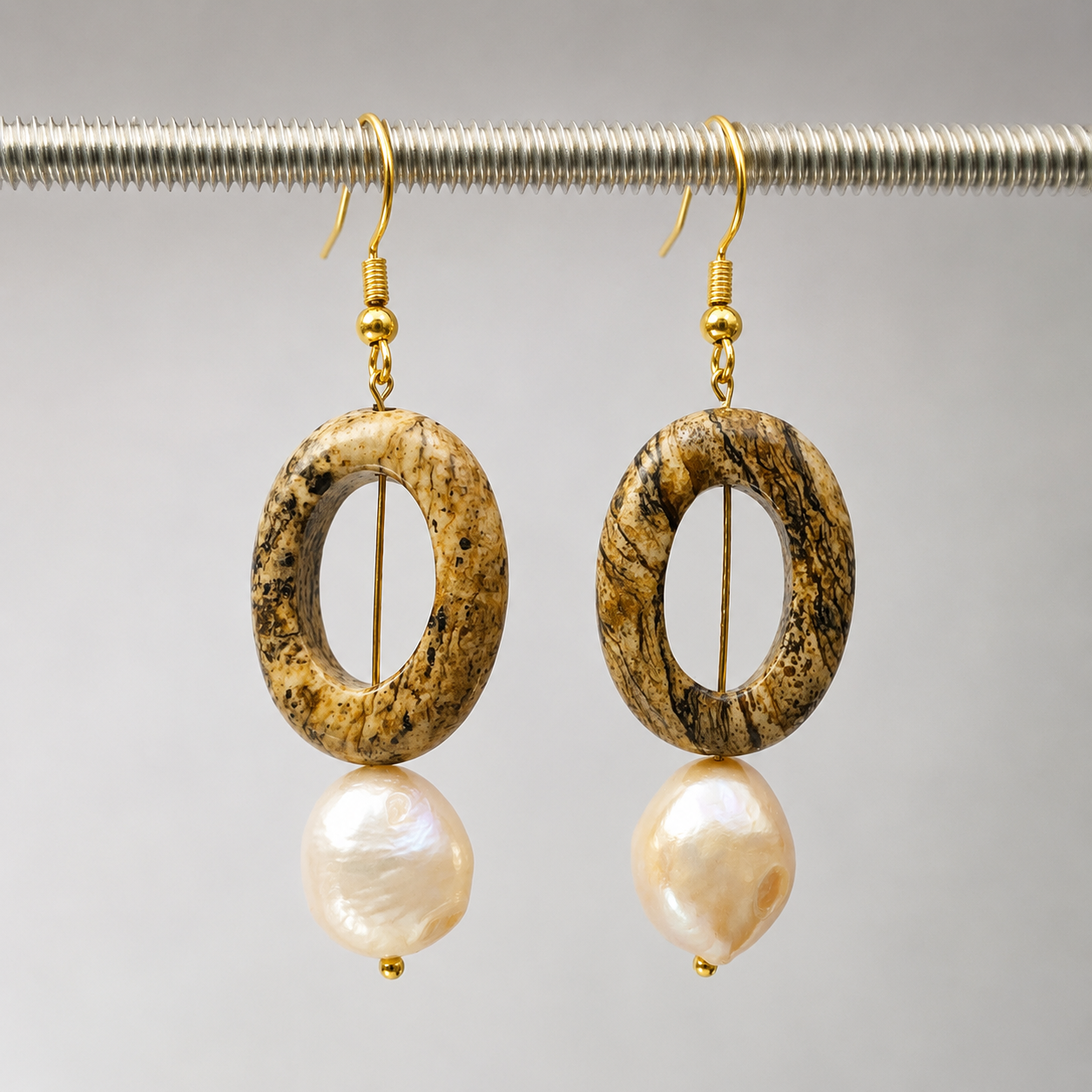 A pair of drop earrings with oval textured brown and beige stone pendants and irregular cream-colored pearl-like drops, hanging from gold hooks against a light gray background.