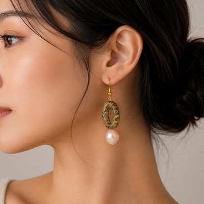 A close-up side profile of a woman wearing a dangling earring with a textured gold oval frame and a white pearl drop, against a soft neutral background.