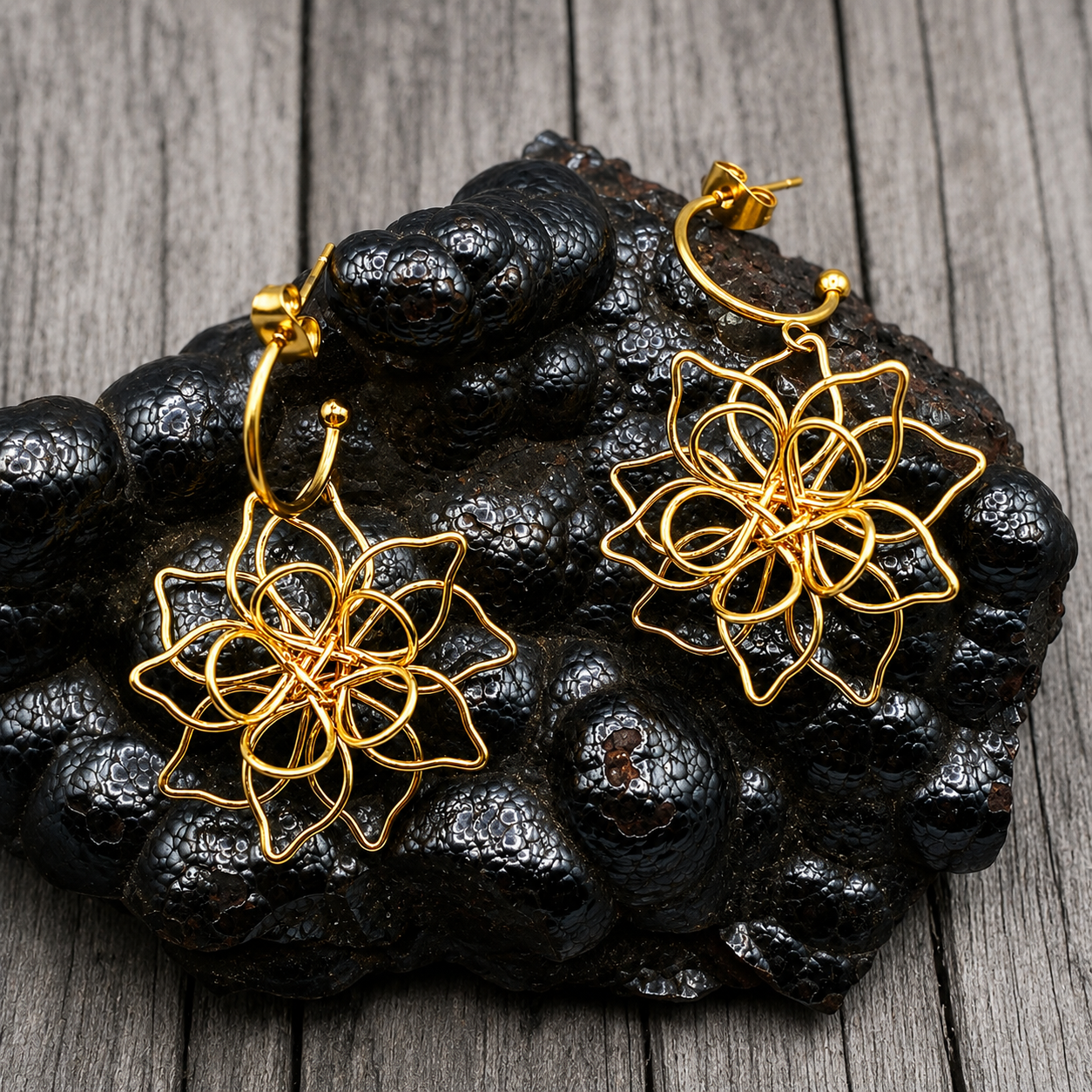 A pair of gold-toned wire hoop earrings with intricate floral or geometric web-like dangling designs, displayed on a dark textured rock against a gray wood plank background.