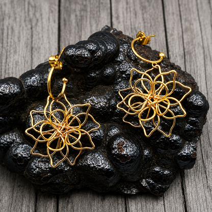 A pair of gold-toned wire hoop earrings with intricate floral or geometric web-like dangling designs, displayed on a dark textured rock against a gray wood plank background.
