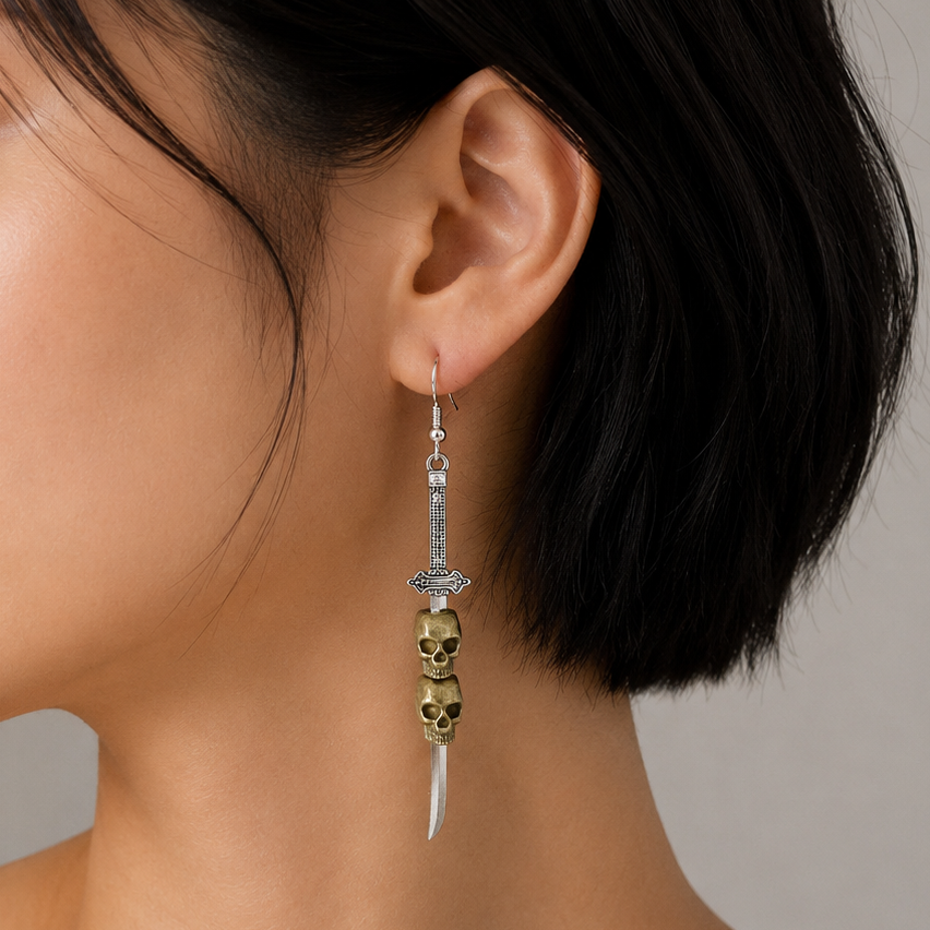 A close-up side profile of a woman with short black hair wearing a dangling earring. The earring features a silver-toned dagger motif with a gold-toned skull accent and a white curved blade tip, set against a neutral gray background.